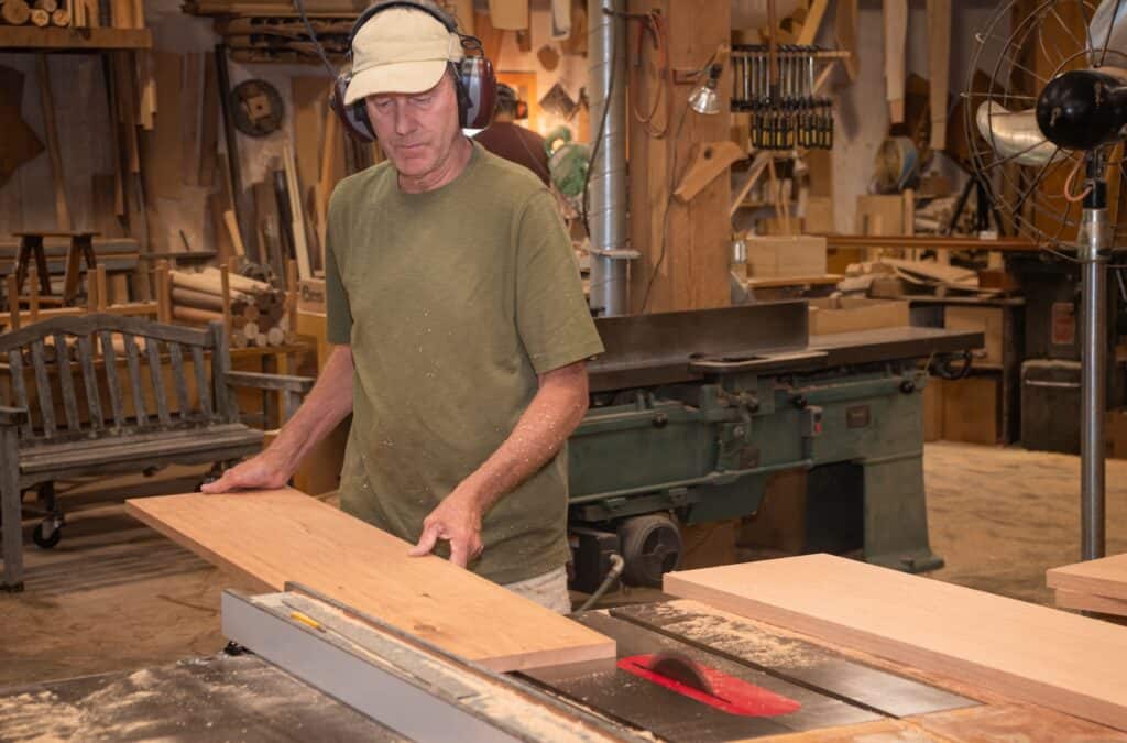 Furniture maker feeding a board through a tablesaw in woodshop.
