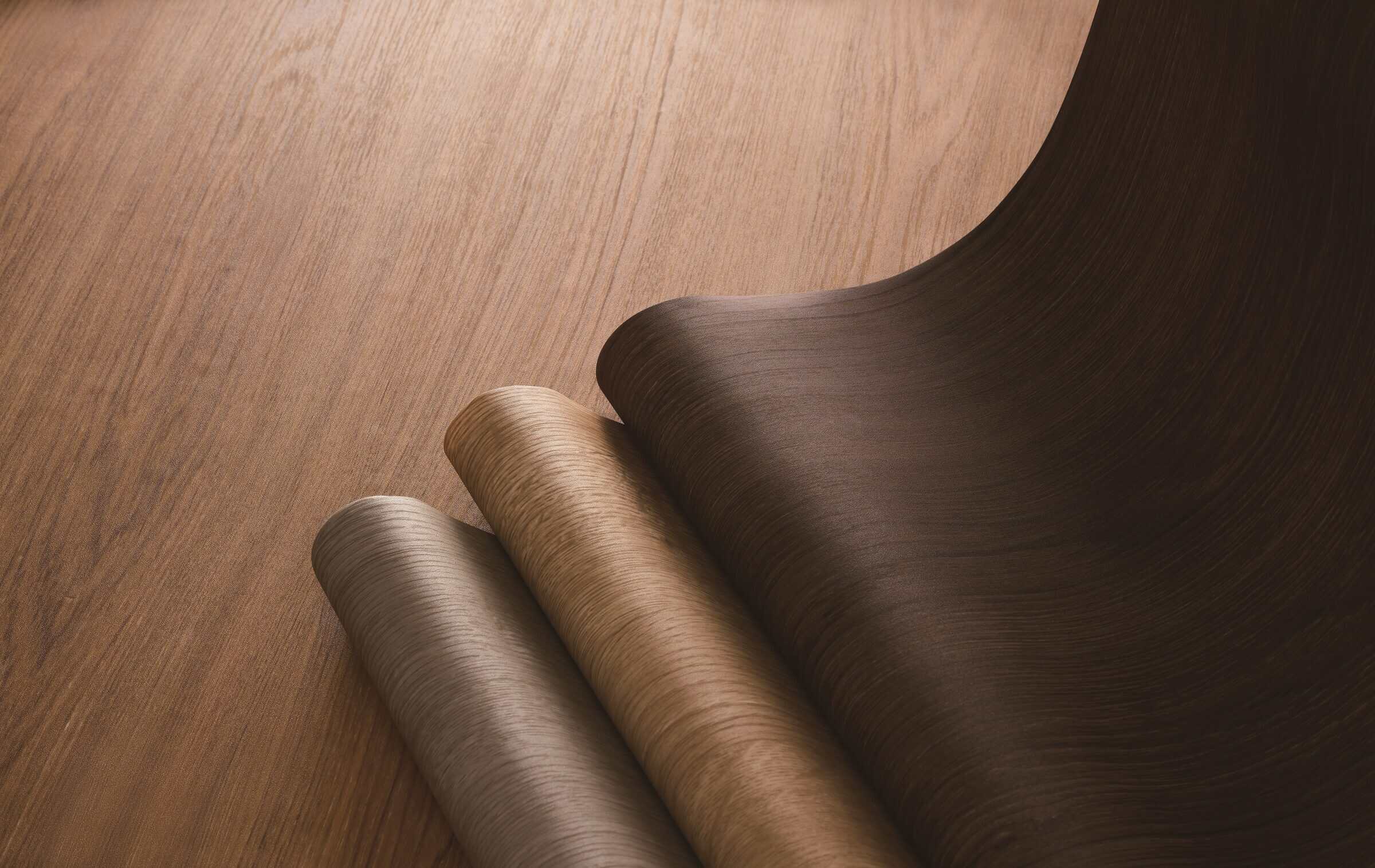 Three partly unrolled rolls of wood-grain architectural film in gray, light oak and dark walnut, resting on a wood-grain surface.