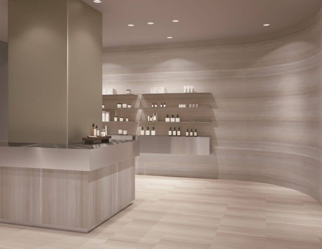 Retail boutique with curved walls, a service counter and display shelving all finished in a pale, linear-grained stone pattern.