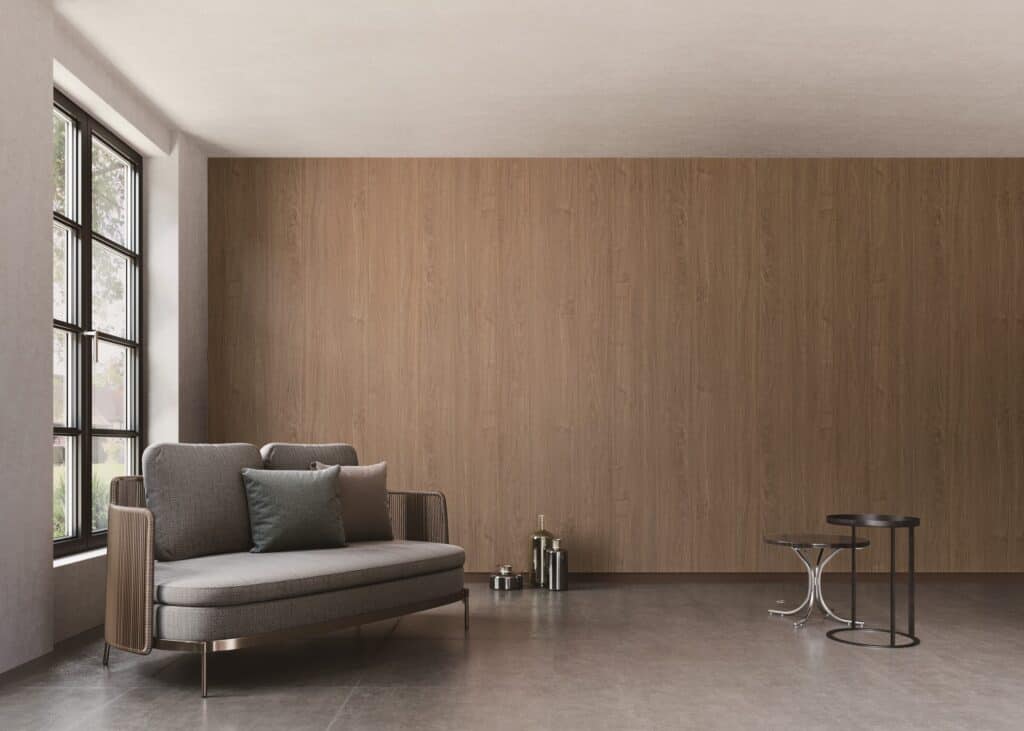 Living room with a full-height warm walnut wood-grain feature wall behind a gray loveseat, beside tall black-framed windows.