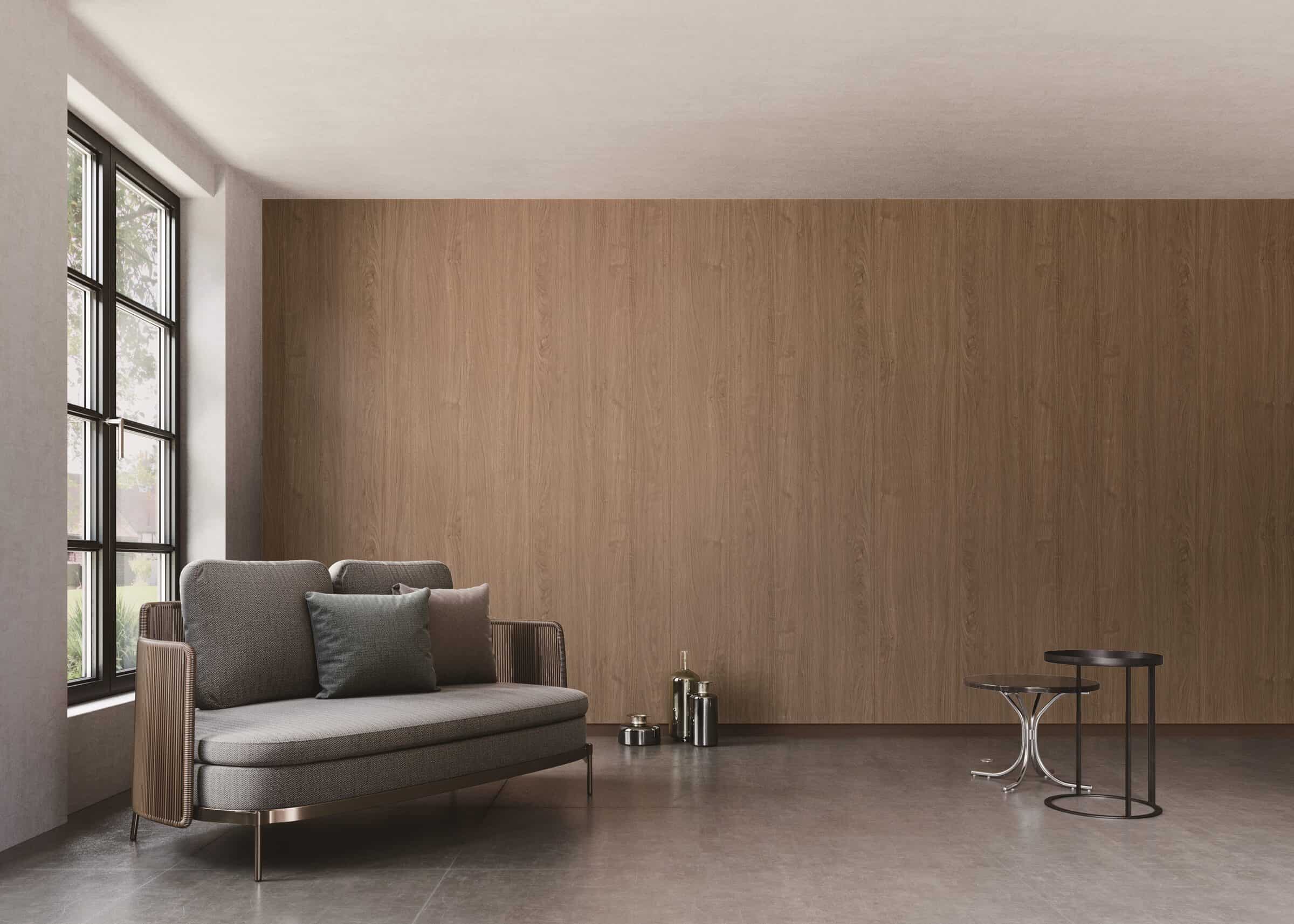 Living room with a full-height warm walnut wood-grain feature wall behind a gray loveseat, beside tall black-framed windows.
