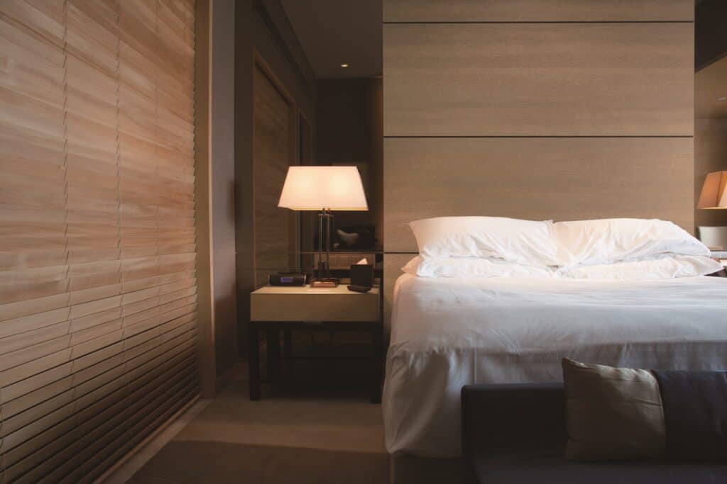 Dimly lit hotel room with a medium brown straight-grain wood headboard wall behind a white bed and bedside lamp.