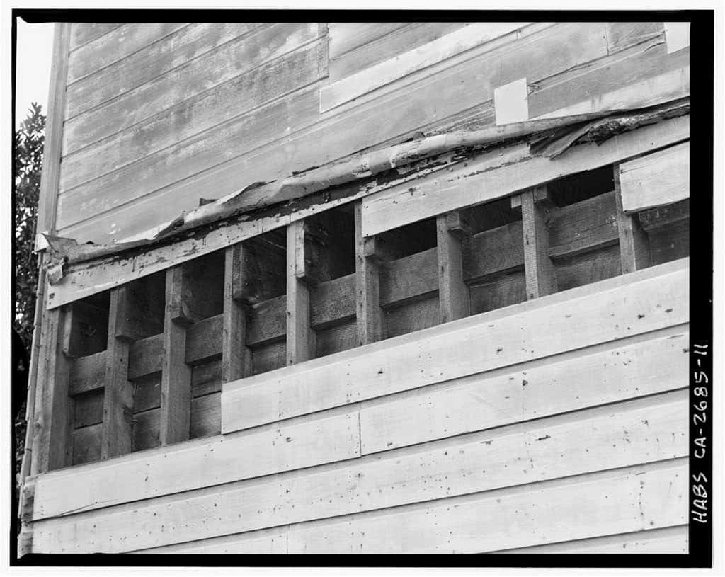 Exposed balloon framing behind removed siding, showing continuous studs notched to carry a horizontal girt that supports second-floor joists