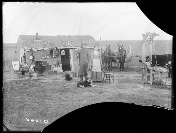 A man and a woman in front of the John Curry sod house, Nebraska, 1886, with a chair, birdcage, antlers, wagon and well frame